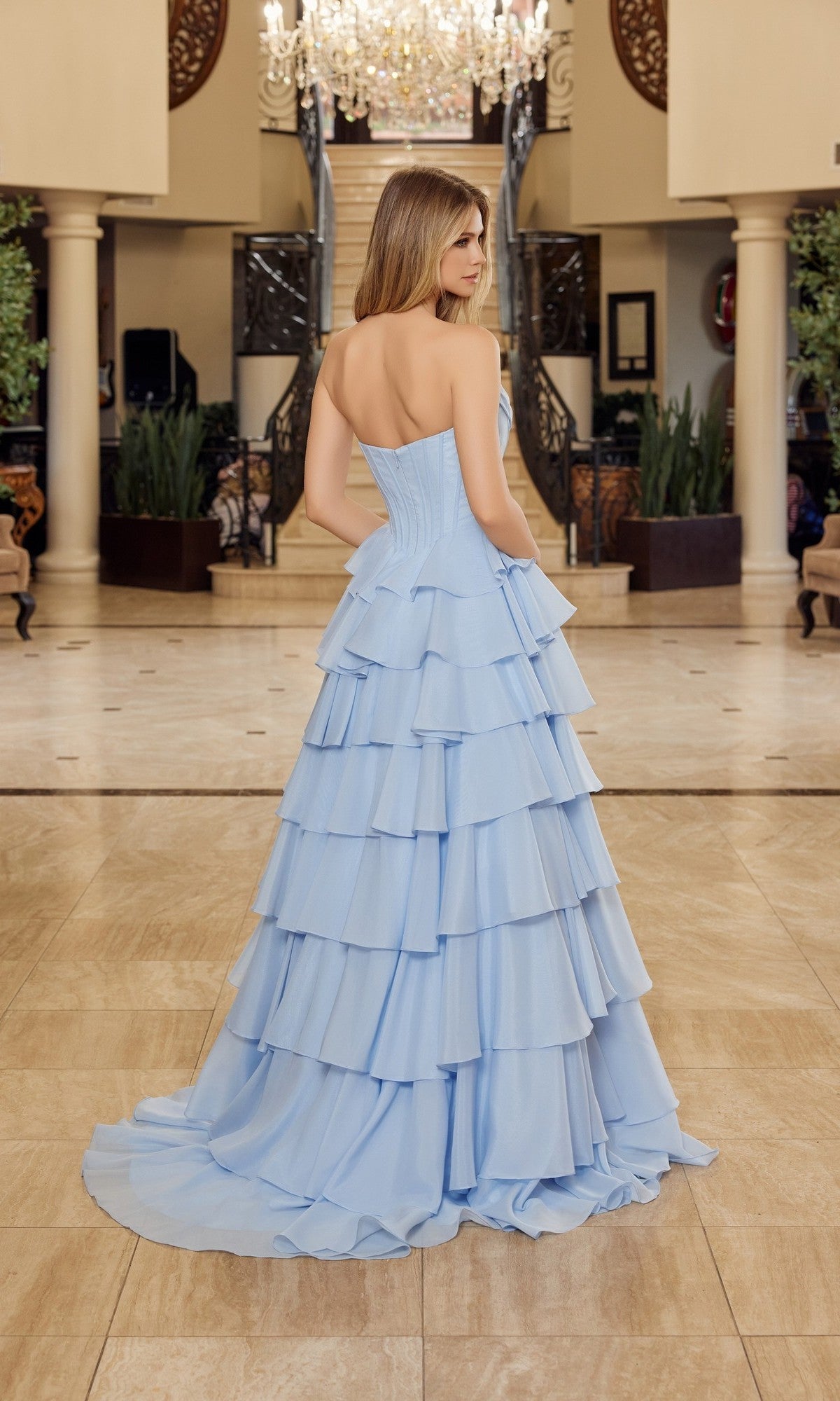 A woman wearing the Nox Anabel Y1806 long prom dress, featuring a strapless bodice and ruffled tiered skirt, stands facing away in an elegant foyer with a staircase in the background.